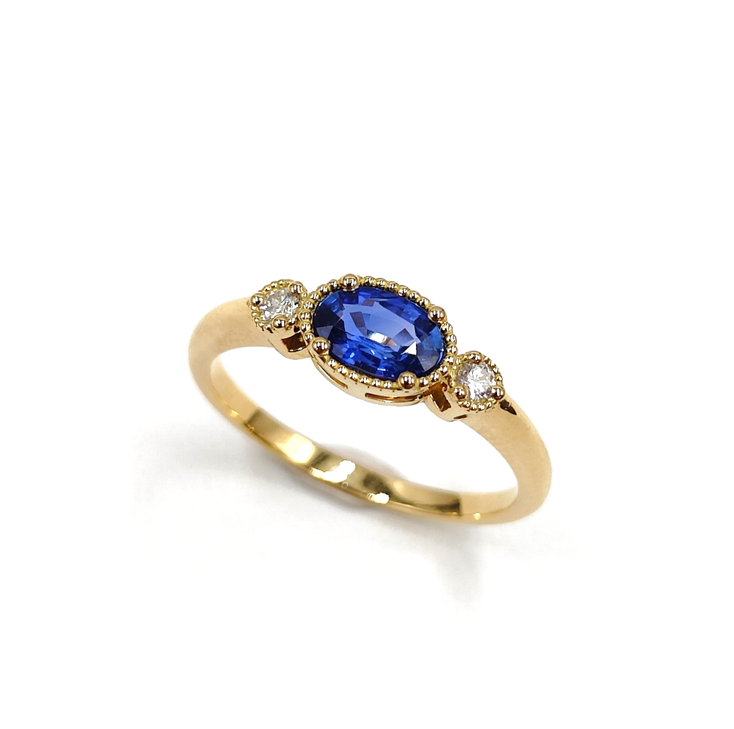 une bague en or jaune sertie d'un saphir bleu ovale central et de deux petits diamants de chaque côté du saphir. Le saphir est entouré d'un fin motif orné sur la monture. La bague est simple et élégante.