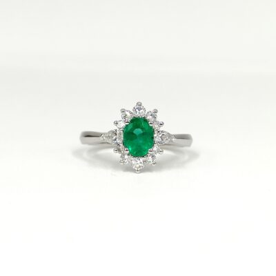 une bague en or blanc sertie d'une émeraude centrale ovale et entourée d'une couronne de diamants taille brillant et taille poire. L'émeraude est d'un vert vif intense. La monture est délicate et élégante.