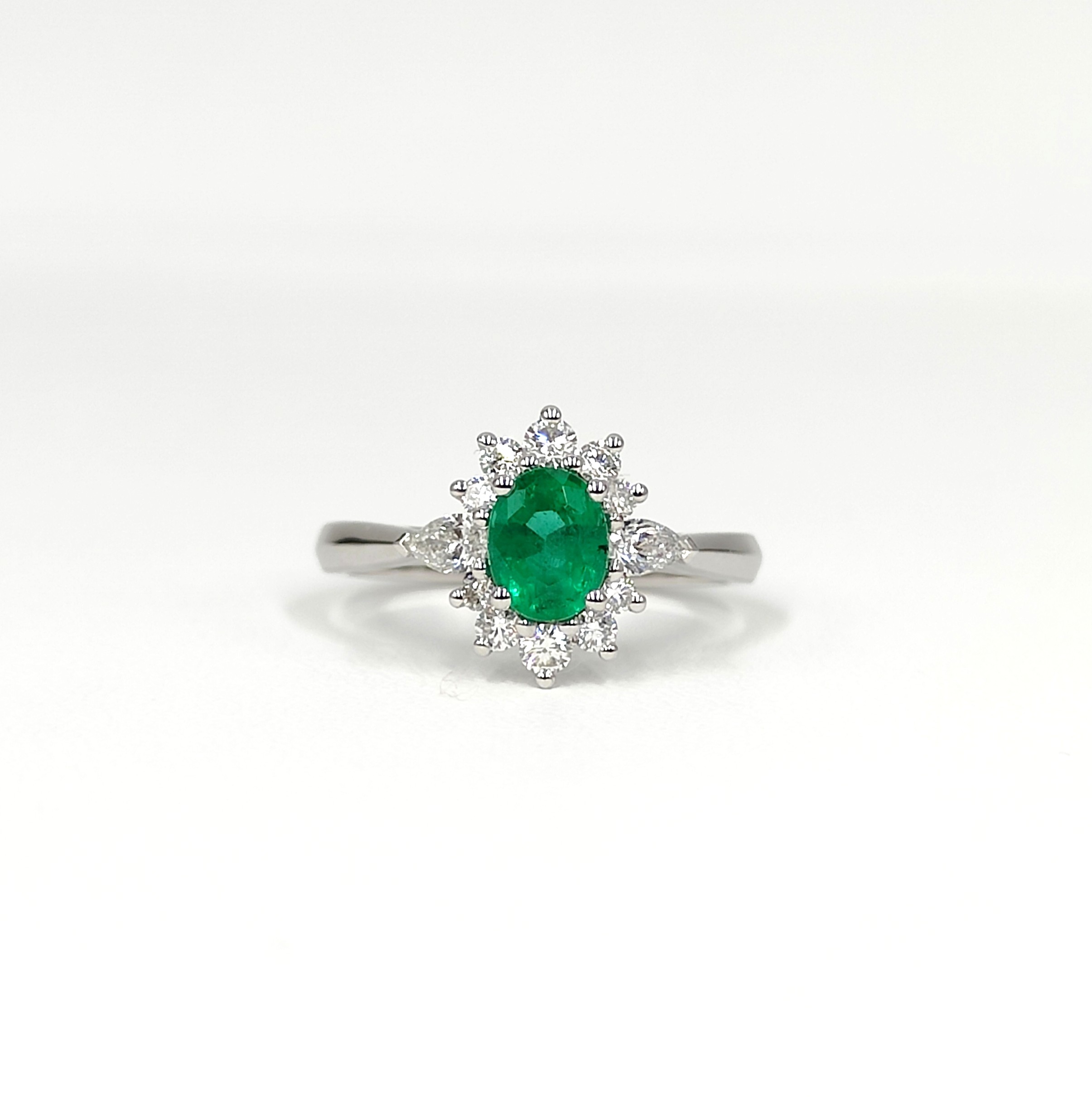 une bague en or blanc sertie d'une émeraude centrale ovale et entourée d'une couronne de diamants taille brillant et taille poire. L'émeraude est d'un vert vif intense. La monture est délicate et élégante.